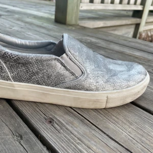 Silvriga slip-on sneakers från Duffy - Snygga silvriga slip-on sneakers från Duffy med en glittrig yta. Skorna har en bekväm passform och är perfekta för en stilren look. De har en vit sula och elastiska paneler för enkel på- och avtagning💓