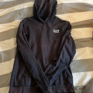 Svart hoodie från Emporio Armani - Säljer en stilren svart hoodie från Emporio Armani med EA7-logga på bröstet. Perfekt för en avslappnad look. Den har en klassisk huva och långa ärmar. Passar bra till både jeans och joggers.