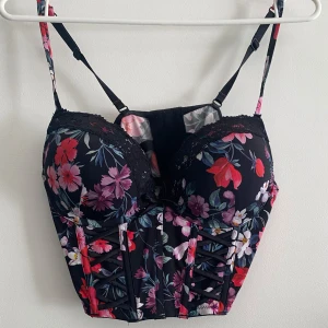 Blommig korsettopp med spetsdetaljer - Snygg blommig korsettopp med justerbara axelband och spetsdetaljer. Toppen har en vacker blommönster i rött, rosa och lila på en svart bakgrund. Perfekt för att ge en extra touch till din outfit.