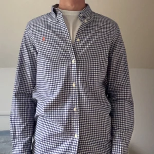 Rutig Ralph Lauren skjorta - Snygg blåvit rutig skjorta från Ralph Lauren med klassisk button-down krage och broderad logga i orange på bröstet. Skjortan är långärmad, har normal passform och är tillverkad i mjuk bomull. Perfekt till jeans eller chinos för en clean look. Ny 2000kr