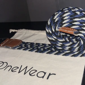 Blåvit flätad OneWear-bälte - Snyggt flätat bälte från OneWear i blått, vitt och svart med bruna skinn-detaljer vid spännet. Bältet har ett silverfärgat metallspänne som lyfter upp hela bältet! Märket är gjort av onewear som drivs av Bruce Lee. Bältet är i bra skick och kan lyfta en outfit. Det passar till alla byxor, t.ex chinos eller ett par stiliga jeans!👏Denna färgen säljs inte längre på onewear!