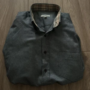 Grå Burberry skjorta limited edition  - Snygg grå skjorta från Burberry med klassiskt rutigt mönster på insidan av kragen och ärmsluten. Skjortan har bröstficka med diskret broderad logga och knäppning framtill. Perfekt för dig som gillar stilrena plagg med en twist.