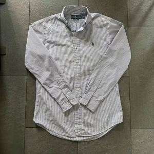 Randig Ralph Lauren Skjorta - blå/vit (Stl S) - Säljer denna snygga randiga skjorta från Ralph Lauren. Skjortan är i gott skick men har en knappt synlig fläck på ena axeln som kanske går bort i tvätt. Nypris ligger på ca 1600kr