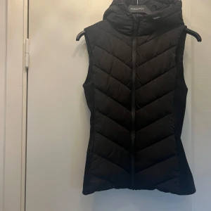 Svart dunväst från KappAhl med huva - Snygg svart dunväst från Zara med quiltad design och dragkedja framtill. Västen har en stor huva och är figursydd för en modern look. Perfekt att slänga över en hoodie eller tröja när det är kyligt ute.