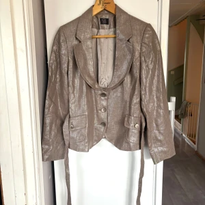 Glansig beige kavaj från GR Design vintage - Snygg bronsig kavaj från GR Design, vintage  med glansig finish och markerad krage. Kavajen har knappar framtill, två fickor med lock och ett bälte i midjan för extra form. Perfekt för dig som vill sticka ut med en unik och trendig look.