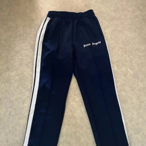 Palm Angels blå trackpants med vita ränder - Mörkblå trackpants från Palm Angels med vita sidoränder och svart detalj, elastisk midja med snörning och dragkedja vid benslut. Snygg logga på framsidan och sportig vibe. Perfekta för en chill och trendig look.