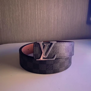 Läder bälte  - Snyggt svart bälte med klassiskt rutigt Damier-mönster och stor logga i silver på spännet. Bältet är gjort i exklusivt skinn och har en lyxig känsla. Perfekt accessoar för att lyfta din outfit med en ikonisk detalj. Den är 110cm. Kommer med en hålslagare.