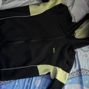 Svart vindjacka från BOSS med huva - Svart vindjacka från BOSS med ljusgula paneler på ärmar och axlar. Jackan har huva med dragsko, hel dragkedja framtill och diskret BOSS-logga på bröstet. Perfekt för dig som gillar sportig och clean stil.