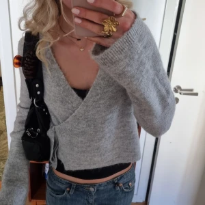 Grå omlott kofta i stickad ull - Knappt använd!! Supermysig grå omlott kofta i stickad ull med knyt i sidan och djup v-ringning. Koftan är croppad och har långa ärmar, perfekt att slänga över ett linne eller topp. Enkel och stilren, passar till jeans eller kjol.