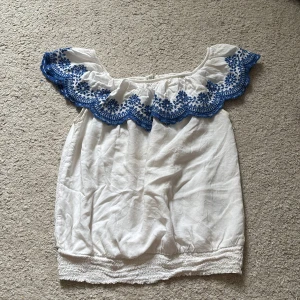 Vit offshoulder blus med blå brodyr H&M - Superfin vit offshoulder blus från H&M med bred volangkant och blå broderade blommor längs axlarna. Blusen har resår nertill och är perfekt för sommarens varma dagar. Luftig och lätt i bomull, passar dig som gillar boho-vibe och snygga detaljer.