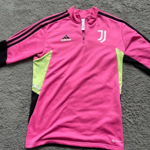 Juventus rosa långärmad träningströja - Juventus långärmad träningströja från Adidas i stark rosa med svarta och neongula detaljer. Tröjan har halv dragkedja, klassiska Adidas-ränder på ärmarna och Juventus-logga på bröstet. Tillverkad i funktionsmaterial (AEROREADY) som andas.
