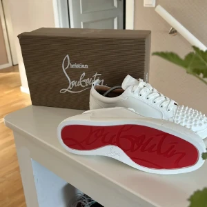 Vita sneakers från Christian Louboutin - Snygga vita sneakers från Christian Louboutin med ikonisk röd sula och coola nitar över tån. Skorna har snörning och är tillverkade i skinn för en lyxig känsla. Perfekta för dig som vill sticka ut med en edgy och exklusiv look. 
