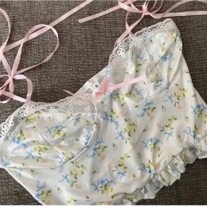 Blommig linnetopp med spets och rosett - Supersöt linnetopp med blommigt mönster i blått, gult och rosa på vit botten. Toppen har tunna rosa knytband, spetskant upptill och en liten rosett framtill. 