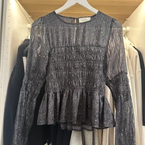 Mönstrad blus från Neo Noir, stl 34 - Supersnygg svart blus från Neo Noir med diskret prickigt mönster i rosa och beige. Blusen har långärmade transparenta ärmar, smockad midja och volang nertill. Tillverkad i tunn, luftig polyester som ger en lätt och flowy känsla.