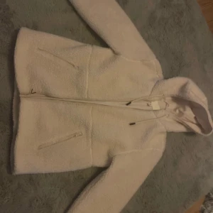 Vit teddyjacka med huva och dragkedja - Mysig vit teddyjacka med huva och dragsko, hel dragkedja framtill och två snedställda fickor med dragkedja. Jackan har lång ärm och är perfekt för kyliga dagar. Oversized och fluffig look som är supertrendig just nu.