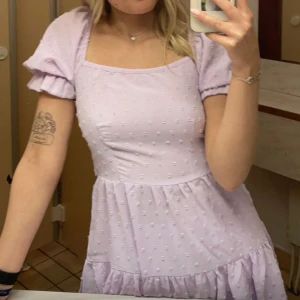 Ljuslila kortklänning med rosetter - Supersöt ljuslila kortklänning med puffärmar, volangkant och strukturmönster. Klänningen har öppen rygg med två stora knytrosetter och fyrkantig ringning. 