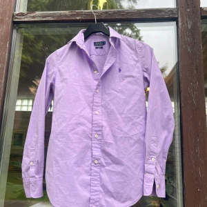 Lila skjorta från Polo Ralph Lauren - Snygg lila skjorta från Polo Ralph Lauren i slim fit-modell. Skjortan har klassisk krage, långa ärmar och knäppning framtill med vita knappar. Broderad logga i lila på bröstet. Perfekt för dig som gillar stilrena och färgglada plagg.