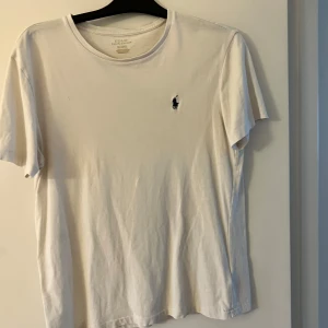 Vit t-shirt från Polo Ralph Lauren - Klassisk vit t-shirt från Polo Ralph Lauren med rund hals och korta ärmar. T-shirten har den ikoniska broderade loggan i mörkblått på bröstet och är tillverkad i mjuk bomull. Perfekt basic-plagg med stilren design.