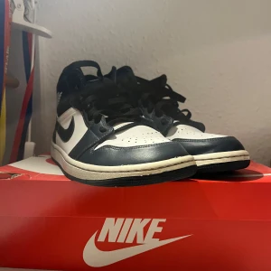 Nike Air Jordan 1 Mid vit/marinblå - Säljer ett par Nike Air Jordan 1 Mid i vit och marinblå färg med klassisk svart swoosh. Skorna har snörning, rund tå och är tillverkade i skinn. Perfekta för dig som vill ha en ikonisk sneaker med stilren vibe och hög komfort.