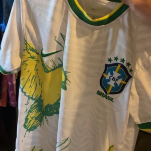 Vit Brasilien fotbollströja Nike - Vit Brasilien fotbollströja från Nike med gröna och gula detaljer, CBF-emblem på bröstet och ett stort grafiskt mönster i gult och grönt. Tröjan har grönt och gult runt kragen och ärmarna. Perfekt för dig som älskar fotboll och vill sticka ut.