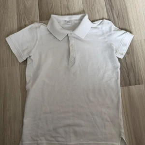 Vit pikétröja från Lindex barn - Klassisk vit pikétröja från Lindex i återvunnen bomull. Tröjan har korta ärmar, krage och knäppning med två knappar framtill. Perfekt basic till garderoben och enkel att matcha med allt.