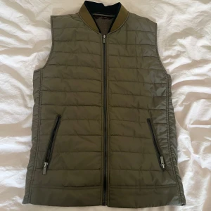 Olivgrön dunväst från Zara Man - Snygg olivgrön dunväst från Zara Man med quiltad framsida och slät baksida. Västen har två dragkedjefickor, ribbad krage och hel dragkedja framtill. Perfekt för lager-på-lager och enkel att matcha med hoodies eller tröjor.