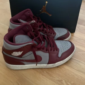 Nike Air Jordan 1 vinröd och grå - Säljer ett par Nike Air Jordan 1 sneakers i en snygg kombination av vinrött och grått. Skorna har klassisk high-top design, perforerad tå och grå sula. Tillverkade i skinn med vinröda snören och ikoniska Jordan-loggan på sidan.