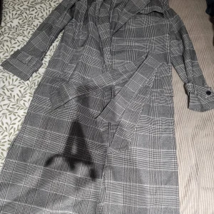 Grå rutig trenchcoat med bälte - Snygg grå trenchcoat med klassiskt rutigt mönster och bälte i midjan. Jackan har långa ärmar med knappar vid ärmslut och en stilren krage. Perfekt för dig som vill ha en tidlös och trendig look.