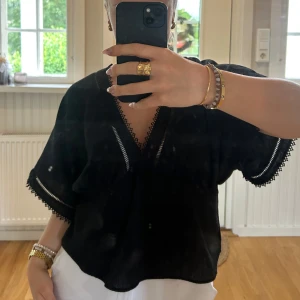 Svart blus med spets från Zara - Svart trendig blus från Zara med v-ringning och fina spetsdetaljer längs halsringning och ärmslut. Blusen har korta, vida ärmar och är tillverkad i ett lätt, luftigt material som känns perfekt för varmare dagar. Storlek M men passar även en S.💖💖