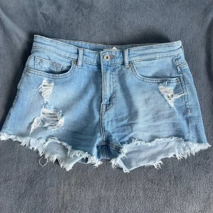 Ljusblå jeansshorts med slitningar - Snygga ljusblå jeansshorts med slitna detaljer och fransig kant. Klassisk femficksmodell med knapp och dragkedja framtill. Perfekta för sommaren och ger en avslappnad känsla.
