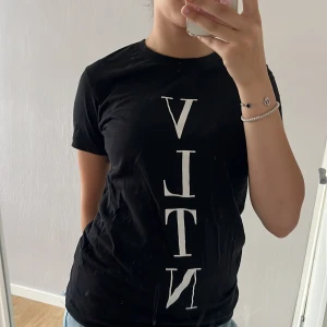 Svart VLTN t-shirt från Valentino - Svart t-shirt från Valentino med stor vit VLTN-logga tryckt vertikalt på framsidan. Klassisk rund halsringning och korta ärmar. Skön passform och stilren design som passar perfekt till jeans eller shorts.