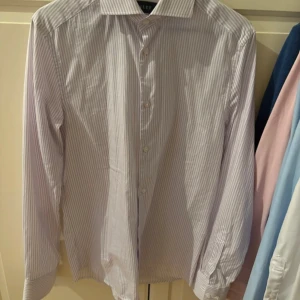 Ljuslila randig skjorta från Riley - Säljer en slim fit skjorta från Riley i ljuslila och vitt med smala ränder. Skjortan har klassisk krage, långa ärmar med dubbla knappar vid manschetten och knäppning framtill. Perfekt för dig som gillar stilrena och fräscha plagg.