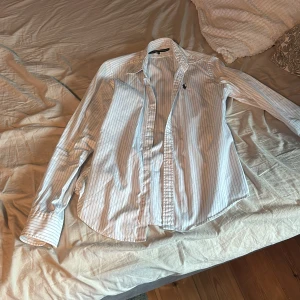 Vitblå randig skjorta Ralph Lauren - Vit skjorta med tunna blå ränder från Ralph Lauren Sport. Klassisk krage, långärmad och med knappar framtill. Broderad logga på bröstet. Skjortan är i bomull och har en tidlös, stilren look som passar perfekt till jeans eller chinos.