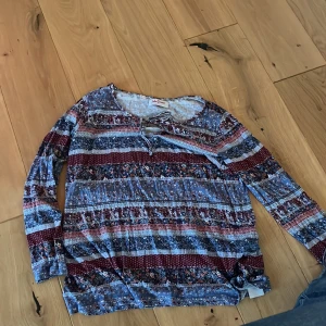 Mönstrad långärmad blus från John Baner - Säljer en långärmad blus från John Baner med snyggt mönster i blått, vinrött och vitt. Blusen har snörning vid halsen och är tillverkad i mjuk bomull. Perfekt för dig som gillar boho-vibbar och färgglada detaljer.