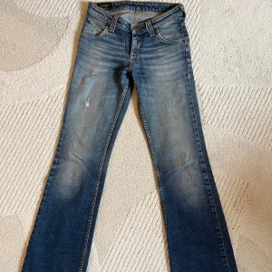Blå lågmidjade bootcut jeans från Lee - Snygga blå jeans från Lee med bootcut passform och klassisk femficksdesign. Jeansen har slitningar och en liten reva på låret för en cool, använd look. Tillverkade i mjukt denim med lätt tvättad effekt och kontrastsömmar.