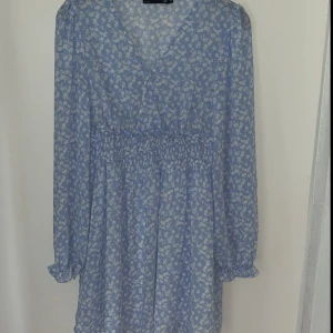 Blå blommig kortklänning med v-ringning - Supersöt blå kortklänning med vita små blommor, v-ringning och långa ärmar med resår vid handlederna. Klänningen har smock i midjan som ger en snygg passform och är gjord i ett lätt, luftigt material. Dubbel underkjol. Perfekt för vår och sommar! Använd en gång 