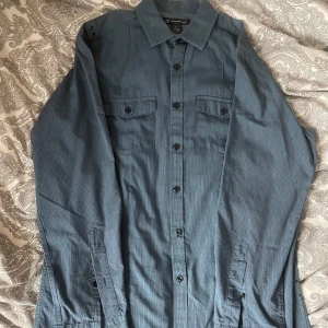 Blå långärmad skjorta - Snygg blå skjorta med diskreta ränder, klassisk krage och svarta knappar hela vägen. Två bröstfickor med lock och knappdetaljer på axlarna ger en cool touch. Perfekt för dig som gillar stilrena plagg med lite edge. Säljer mycket skjortor så köp gärna flera i ett bundle!