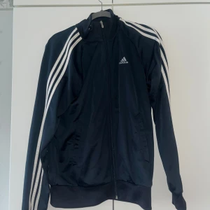 Svart Adidas vindjacka med vita ränder - Klassisk svart Adidas vindjacka med vita ränder längs ärmarna och dragkedja framtill. Jackan har hög krage, två fickor och den ikoniska Adidas-loggan på bröstet. Perfekt för en sportig och avslappnad stil.