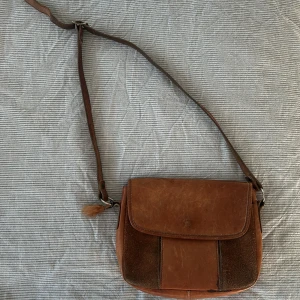 Brun axelväska i skinn med justerbart band - Säljer en brun axelväska i skinn med justerbart axelband och klassiskt spänne i metall. Väskan har flera fack inuti och dragkedja för extra säkerhet. Perfekt storlek för det viktigaste och snygg vintagekänsla💕