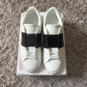 Valentino Garavani Open sneakers vit/svart - Snygga Valentino Garavani Open sneakers i vitt skinn med bred svart läderdetalj över snörningen. Klassisk rund tå, platt sula och vita skosnören. Perfekta för dig som vill ha en clean look. Hör av dig vid frågor och funderingar, kvitto finns 😃