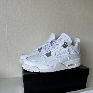 Nike Air Jordan 4 White Metallic Silver - Nike Air Jordan 4 i helvitt med metallic silver-detaljer. Skorna har klassisk meshpanel, snörning och Jumpman-logga i silver på hälen. Ovandel i skinn och mesh ger en clean och modern look. Perfekta för dig som vill sticka ut med stilrena sneakers.