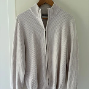 Vit stickad kofta från EAST WEST - Säljer en stilren vit stickad kofta från EAST WEST i storlek M. Koftan har hel dragkedja framtill, hög krage och långärmad passform. 