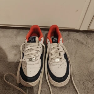 Nike Air Force 1 i vitt, blått och rött - Snygga Nike Air Force 1 sneakers med vit bas, mörkblå detaljer i jeansmaterial och röd textil runt ankeln. Klassisk design med perforerad tå, vita snören och tydlig Nike-logga på sidan. Perfekta för dig som vill sticka ut med en unik färgkombo. Storlek 39