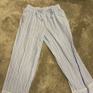 Randiga pyjamasbyxor från Hunkemöller - Blå-vita pyjamasbyxor från Hunkemöller i storlek S. Byxorna har tunna vita ränder, resår i midjan och är tillverkade i mjuk bomull. Perfekta för chill dagar hemma eller mysiga kvällar. Klassisk look med bredare ben och bekväm passform.