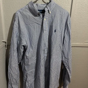 Randig skjorta från Ralph Lauren - Klassisk blåvit randig skjorta från Ralph Lauren med broderad logga på bröstet. Skjortan har lång ärm, knappar framtill och är tillverkad i 100% bomull. Perfekt för dig som gillar stilren och tidlös design.
