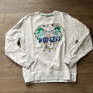 Grå Kenzo sweatshirt med elefant - Snygg grå sweatshirt från Kenzo med stort broderat elefanthuvud och Kenzo Paris-logga på bröstet. Tröjan har rund hals, långa ärmar och är gjord i mjuk bomull. Perfekt statement-plagg för dig som gillar streetstyle och coola prints.