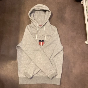 Grå hoodie från GANT med logga - Snygg grå hoodie från GANT med broderad logga och amerikanskt sköldmärke på bröstet. Tröjan har huva med vita snören och en stor magficka. Perfekt för dig som gillar klassisk och sportig stil.