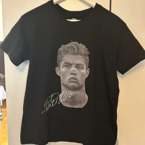 Svart t-shirt med tryck och signatur Ronaldo  - Svart t-shirt i 100% bomull med coolt tryck av ett porträtt och signatur på framsidan. Baksidan har ett stort grafiskt tryck med texten 'CUZ ANYTHING IZ YOURS'. Klassisk passform och rund hals, perfekt för dig som gillar streetstyle. Plagget är helt nytt men på grund av frakten så har den blivit skrynklig.