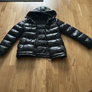 Svart pufferjacka från Alessandro Zavetti - Snygg svart pufferjacka från Alessandro Zavetti med glansig finish och huva. Jackan har dragkedja framtill, två sidofickor med dragkedja och quiltad design. Perfekt för kalla dagar när du vill hålla stilen och värmen.