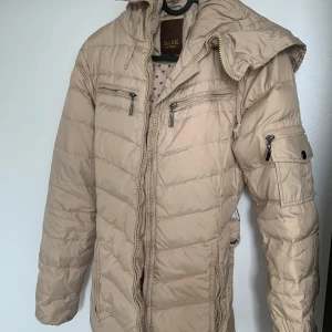 Beige dunjacka från Bark of Sweden - Snygg beige dunjacka från Bark of Sweden med quiltad design, huva och flera dragkedjefickor. Jackan har bälte i midjan och praktisk ficka på ärmen. Perfekt för kalla dagar och riktigt skön att bära.